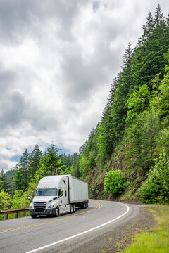 Vertically composed photo with white big rig semi truck and dry van semi trailer running on the mountain road with high rock cliff on the side