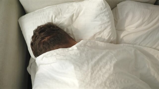 Sleepy shirtless man pulls white duvet over his head to hide from bright morning sun. Top view of tired man burrowing into seersucker pillow on beige sofa bed. Lazy weekend morning and oversleeping.