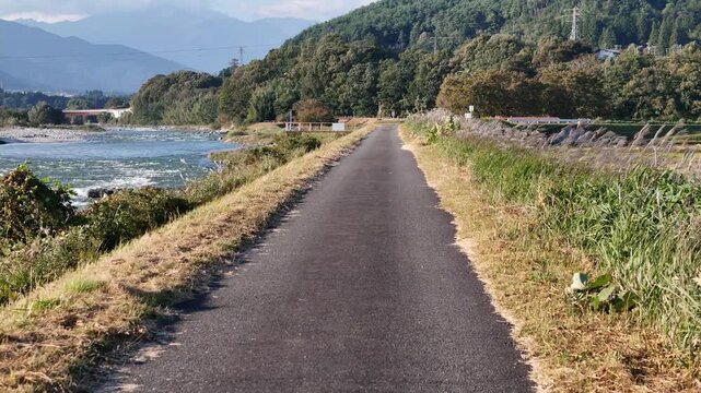 A paved path stretches past grasses and riverside trees with a flowing river on one side and wooded hills beyond.