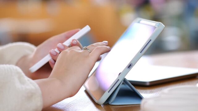 Hands use stylus on tablet. Tablet stands on table. Background is softly blurred