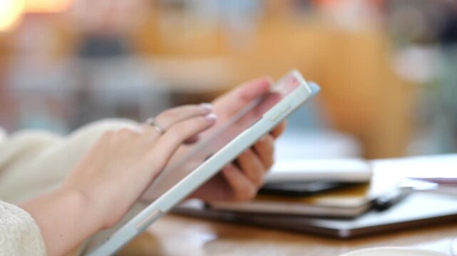 Close-up of hands using a tablet. Fingers swipe screen gently. Background blurred coffee shop setting. Wooden table with notebooks nearby. Warm lighting enhances calm atmosphere