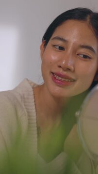Young Asian Woman Skincare Routine in Mirror, Gently Touching Cheek and Face While Adjusting Dark Hair, Calm Beauty Reflection in Soft Natural Light, Relaxed Indoor Portrait