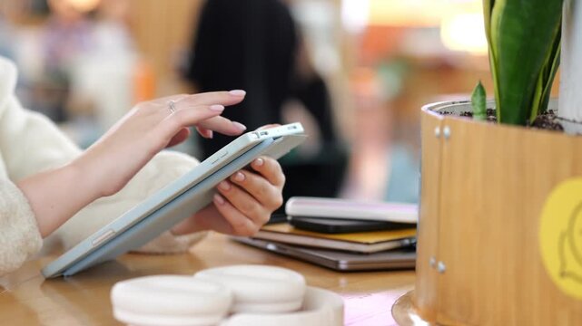 Person using tablet at wooden table. Green plant in wooden pot nearby. Blurred background with people and warm lighting