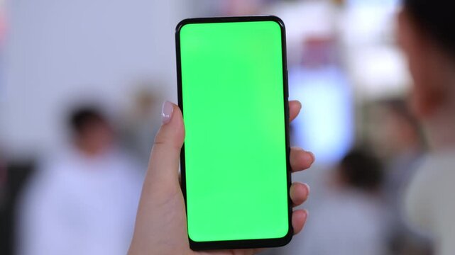 Hand holds smartphone with green screen. Background shows blurred indoor crowd. Focus remains on phone screen only. Person's nails clean and well-groomed. Light reflects off phone edges naturally
