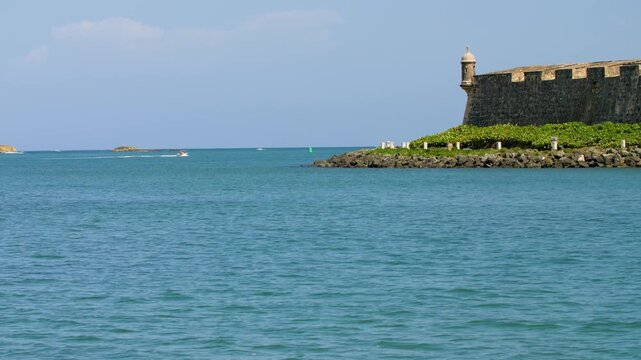 A view of one of the Garita lookouts at Fort San Felipe del Morro in San Juan, Puerto Rico under clear blue skies. Stoned fortified wall construction as small vessel travels along the bay.