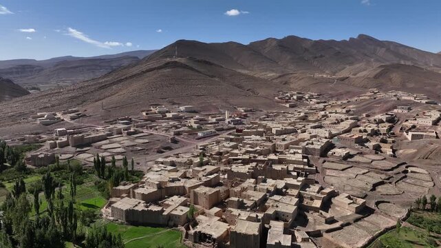 Aerial view of a traditional mountain village with clustered earthen houses, green fields and rugged arid peaks in Morocco's Eastern High Atlas.