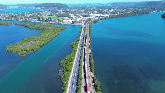 Drone aerial landscape of a freight train carrying shipping containers travelling along railway tracks into the town of Woy Woy alongside Brisbane Water estuary and a bushland reserve in NSW Australia