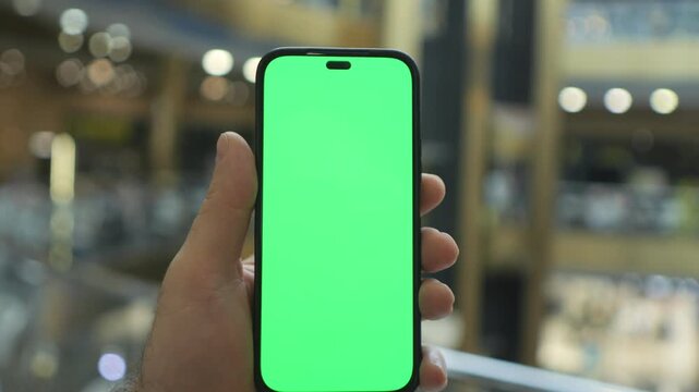 Hand holds smartphone with green screen. Shopping mall background is blurred. Focus on phone display. Hand moves slowly. Mall lights create bokeh effect. Background details visible