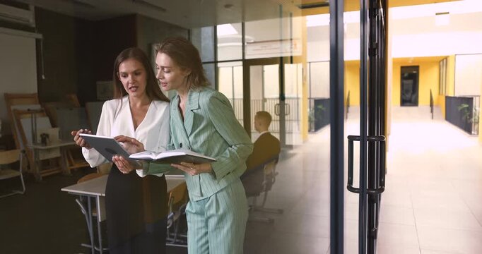 Two businesswomen coworkers engaged in collaborative work with digital tablet and paper notebook in glass office managing tasks reviewing data filling in electronic form in corporate app. Copy space