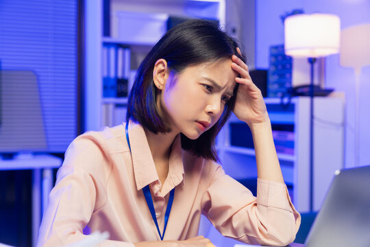 Stressed young Asian office worker feeling overwhelmed with paperwork and workload while working on laptop in office.