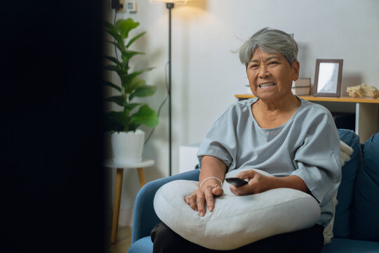 Elderly Asian woman relaxing on a sofa at home, holding a television remote while enjoying a favorite program.