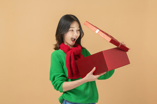 Cheerful Asian woman wearing a green sweater and red scarf, excitedly opening a large red gift box, Christmas celebration, festive holiday spirit.