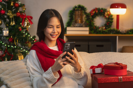 Cheerful Asian young woman wearing a white sweater and red scarf, sitting on a sofa and using her smartphone for online messaging during Christmas, festive holiday.