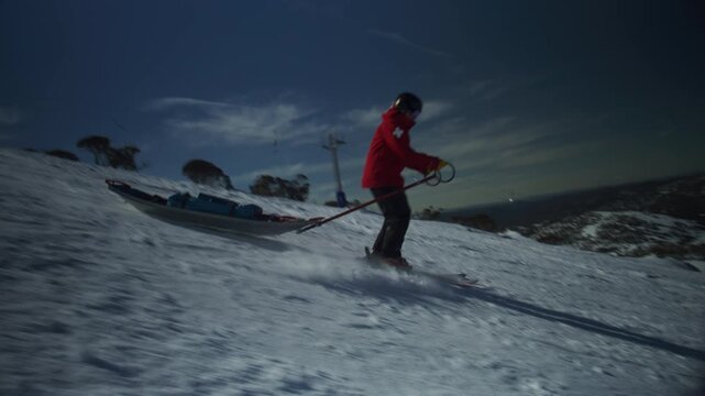 Ski patroller guides a rescue toboggan down a snowy mountain slope beneath a bright blue sky