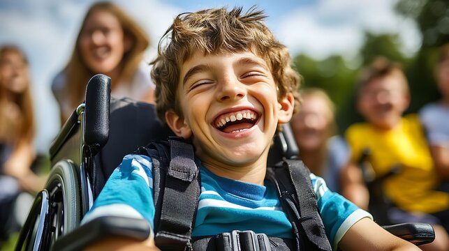 Disabled boy laughing happily sitting in a modern wheelchair surrounded by cheerful friends outdoors during sunny daylight