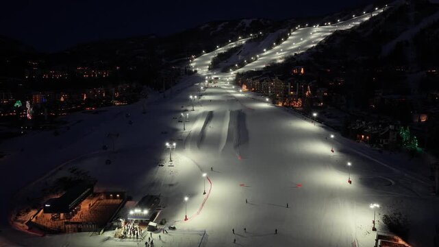 Aerial night skiing in Steamboat Springs, Colorado captures illuminated runs and a snow half pipe, with skiers descending the mountain beside glowing hotels, lifts, and resort lights below.