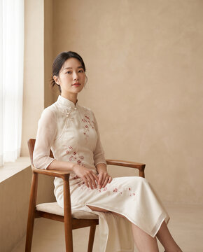 Elegant young Asian woman sitting in modern ivory cheongsam dress  