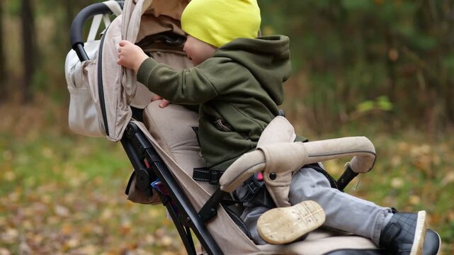Cute Child Explores Stroller Canopy During Baby Outdoor Walk Through Serene Woodland. Curious Infant Turns Around And Smiles From Pushchair Seat On Peaceful Scenic Family Outing In Nature.