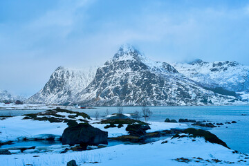 Dramatic winter landscape of Lofoten Islands in Norway. Majestic snow-covered mountain peak towering over cold sea fjord with rocky snow-covered shore in the foreground. Beautiful Arctic nature scener © bank215