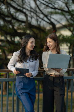 Two Asian businesswomen are discussing a work project while holding documents and a tablet outdoors, near an office building.