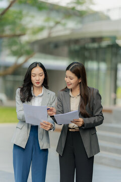 Two Asian businesswomen are discussing a work project while holding documents and a tablet outdoors, near an office building.