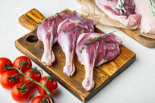 Vine of bright red cherry tomatoes resting beside three raw duck leg thighs garnished with fresh green thyme sprigs on a rustic wooden cutting board against textured white stone surface