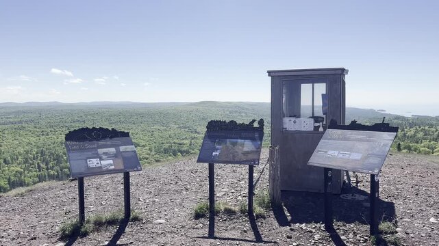 Rightward pan across a stone wall and interpretive displays beside a small booth at Brockway Mountain Overlook in Michigan with green forest stretching into the distance.