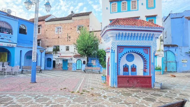 Wide daylight view of the lively cobblestone main square featuring traditional blue-painted arches, cafes, a central fountain pavilion, and the Rif mountains rising behind Chefchaouen