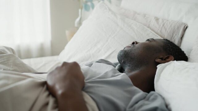 Worried adult black man lying in bed in morning, grabbing blanket, insomnia. Depression, panic attack and intrusive thoughts, middle-aged african american man sleeping in modern hotel room, wake up