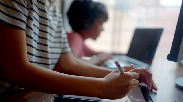 Young student drawing on a digital tablet with a stylus for an e-learning class while his brother studies with a laptop and headphones at home in a bright room
