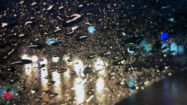 View from inside a moving car through a rain-soaked windscreen at night, with blurred streetlights and glowing light trails creating an atmospheric night time driving scene