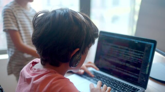Young programmer boy learning coding on laptop, child developing skills with e-learning, focused student typing on keyboard, mother supervising her son studying at home
