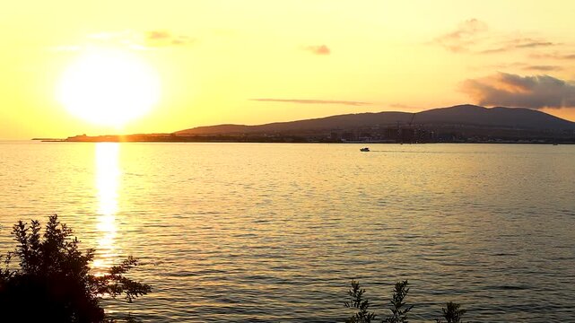 Sunset casts warm hues over water as boats move smoothly, framed by distant hills and lush vegetation, capturing a tranquil coastal atmosphere