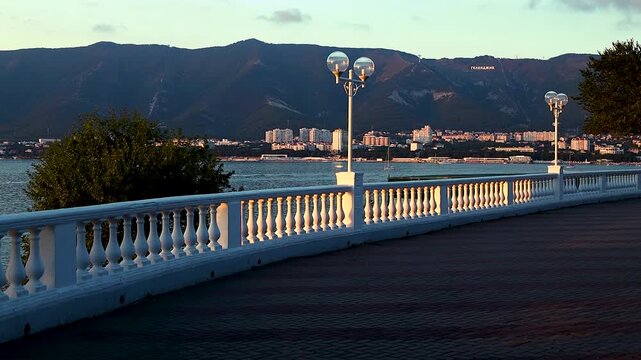Scenic coastal promenade with white railing and street lamps overlooking the bay, mountains in the background, and urban skyline at sunset in a tranquil setting
