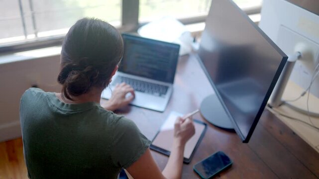 Female programmer coding on a laptop and writing notes on a tablet, a professional developer multitasking at her home office desk during a remote work session