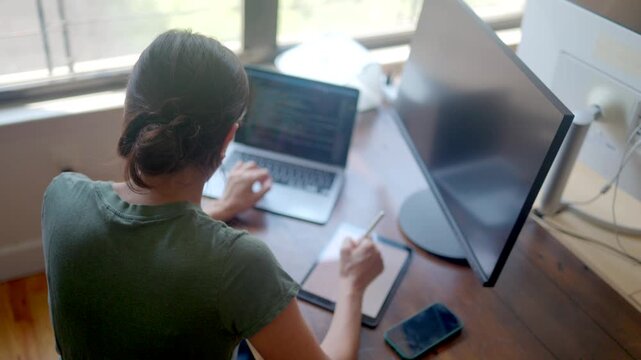 Female programmer and digital artist working from home, coding on a laptop, and drawing on a tablet with a stylus. High angle view of a woman multitasking at her workstation