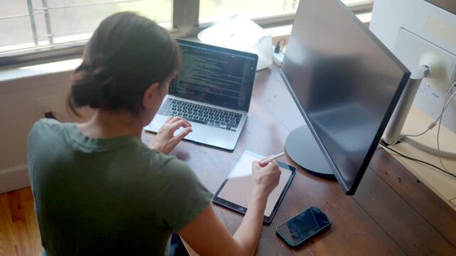 Woman freelancer software developer coding on a laptop and taking notes on a tablet with a stylus. Millennial working from her home office with multiple screens and devices