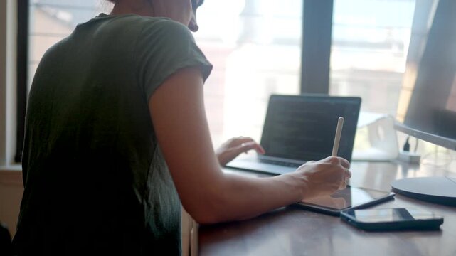 Woman freelancer programmer working from home office, coding on a laptop and using a graphics tablet with stylus, multitasking in a remote workspace with bright window light