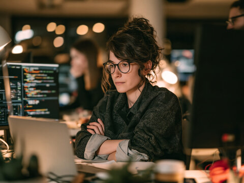 Focused young female programmer working late at night in modern office debugging code on computer screens.