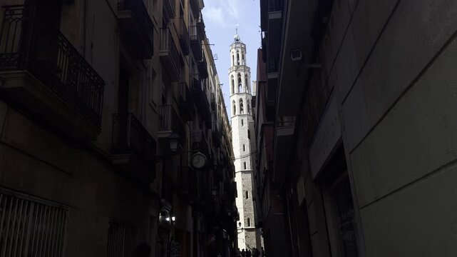 Barcelona, Spain, August 13, 2026. Tall Gothic bell tower of Santa Maria del Pi rising above a narrow medieval street, framed by historic buildings with wrought-iron balconies.