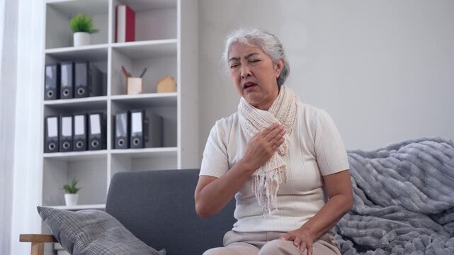 Elderly asian woman with bronchitis coughing and choking while sitting on a sofa at home, feeling unwell with chest pain