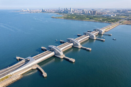 Aerial view of coastal flood defense system with sea barrier, bridge, and distant city protecting urban shoreline from rising water.