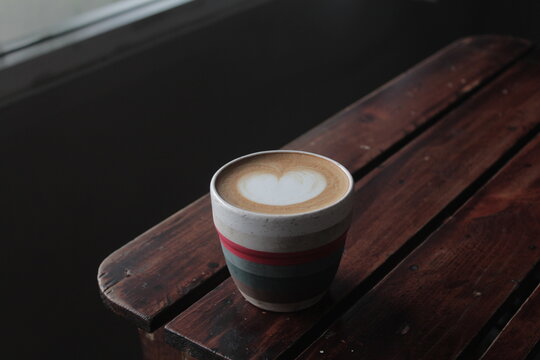 Hot cup of coffee latte with heart latte art served on a dark rustic wooden bench near window light. Cozy and moody aesthetic atmosphere in a cafe, perfect for coffee lover concepts, and menu designs.