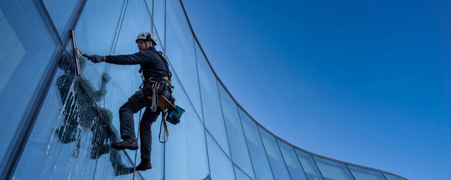 Window cleaner hanging on ropes washing glass facade of skyscraper. Professional male industrial climber working at height. Building maintenance, commercial exterior cleaning service concept.