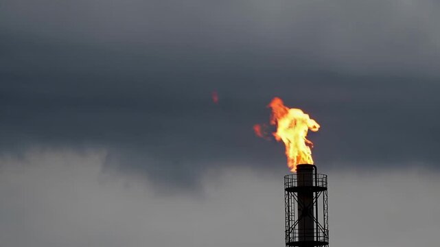 Gas flare stack at a petrochemical refinery burning off excess flammable gases, releasing pollutants and greenhouse gases into the atmosphere against a dramatic, overcast and cloudy sky