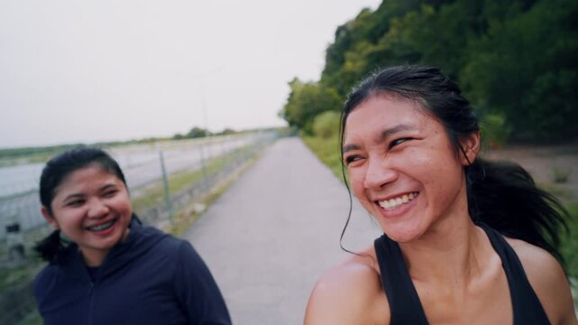 Friends running together, two cheerful young Asian women jogging and laughing on outdoor path