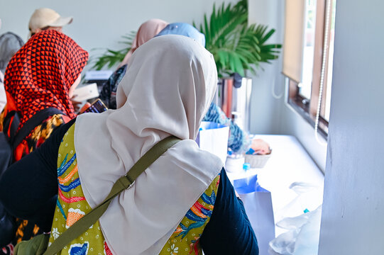 Back view of Muslim women wearing hijabs and traditional batik clothing standing indoors during a workshop, event, or seminar gathering near a table.