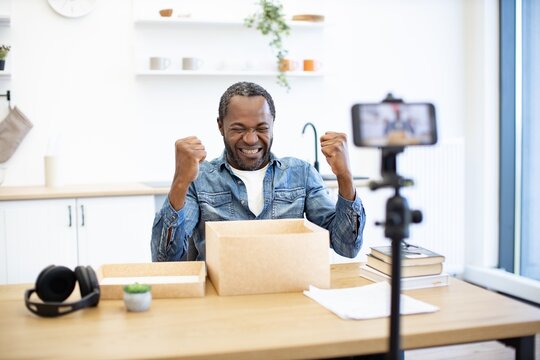 An African American man celebrates at a home office desk with raised fists beside a cardboard box, headphones, books, and a phone on a tripod