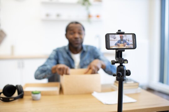 An African American man records a tabletop presentation with a smartphone, tripod, cardboard box, and papers in a bright home workspace