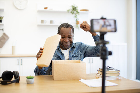 An African American man celebrates while unboxing a package at a home workspace, as a smartphone on a tripod records his smiling reaction
